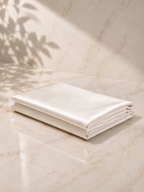 A folded white silk pillowcase set rests on a light beige marble surface with soft sunlight and leaf shadows cast across the scene.