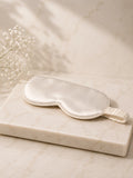 A silk sleep mask in a warm off-white or cream color, resting on a rectangular marble block with delicate baby's breath flowers in the background.