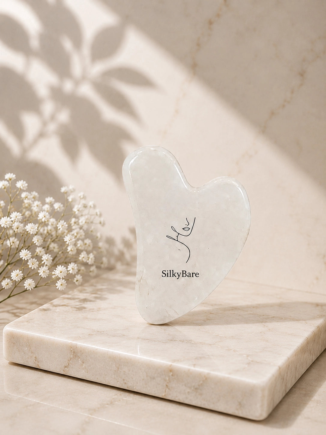 A heart-shaped jade Gua sha massage tool by SilkyBare, standing upright on a polished marble tray. The scene is styled with delicate baby's breath flowers and soft leaf shadows cast on a marble background.