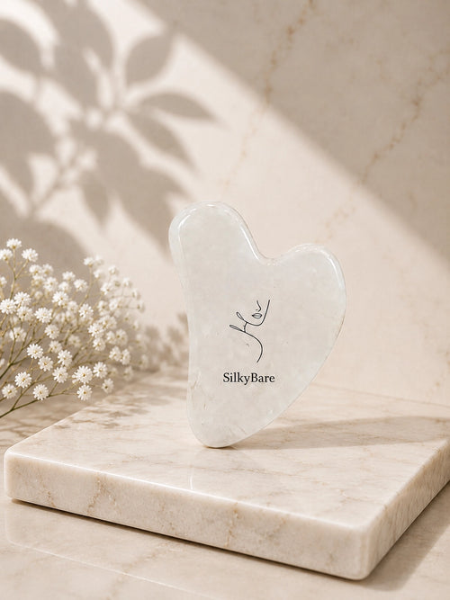 A heart-shaped jade Gua sha massage tool by SilkyBare, standing upright on a polished marble tray. The scene is styled with delicate baby's breath flowers and soft leaf shadows cast on a marble background.